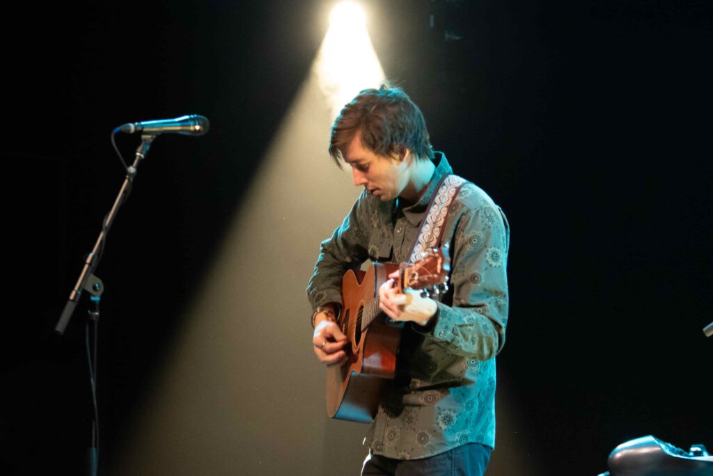 trent performing in spotlight with guitar at microphone